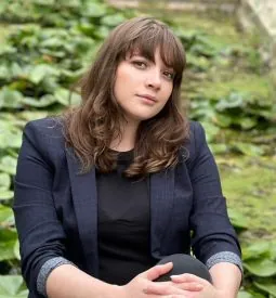 A woman with brunette, slightly curly hair and brown eyes. She wears dark navy blue blazer.